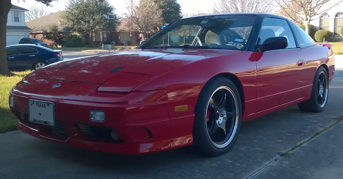 A red Nissan 200SX S14 with black wheels and a Texas license plate is parked on a residential street. The car has a '203'…