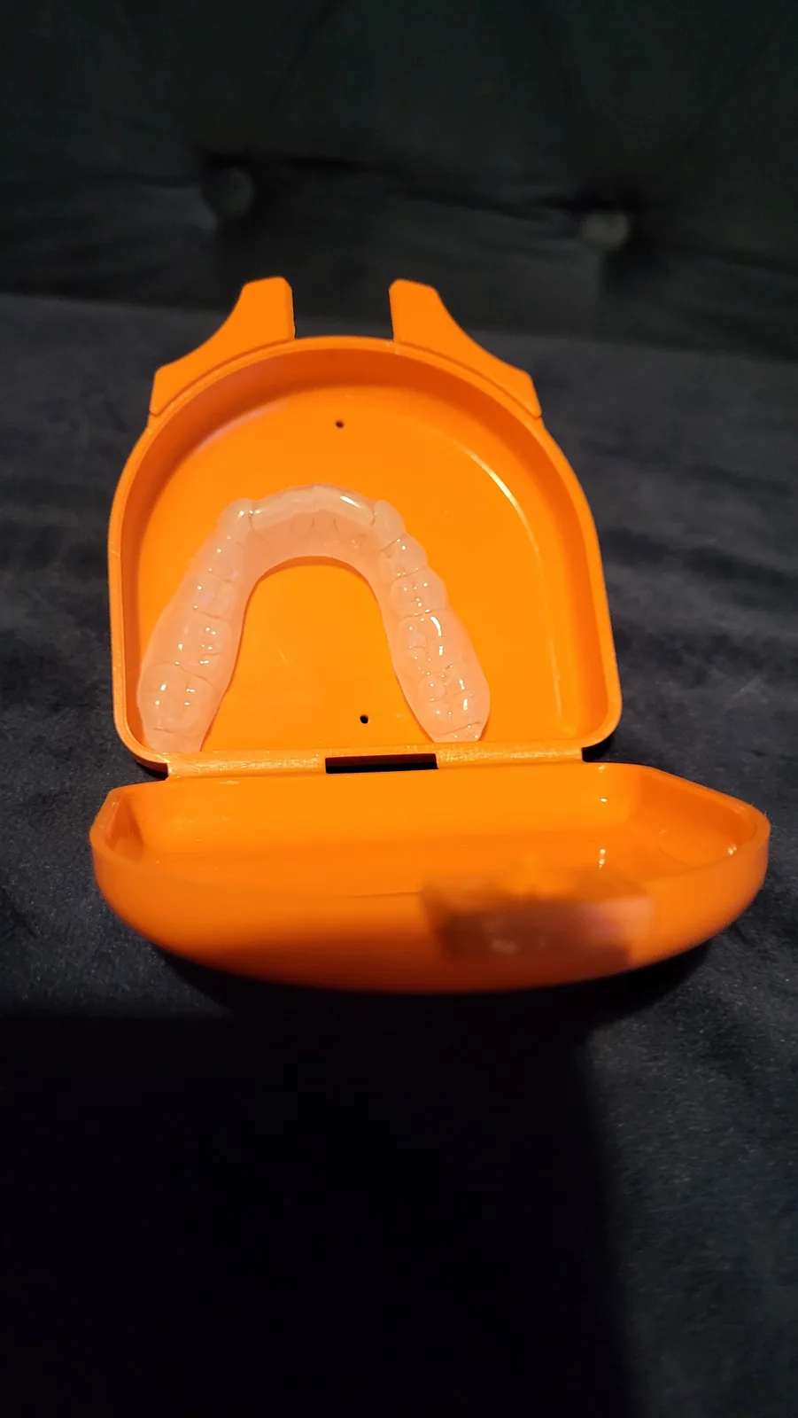 A clear dental aligner rests inside an open, bright orange plastic case, which is sitting on a dark fabric surface. The…