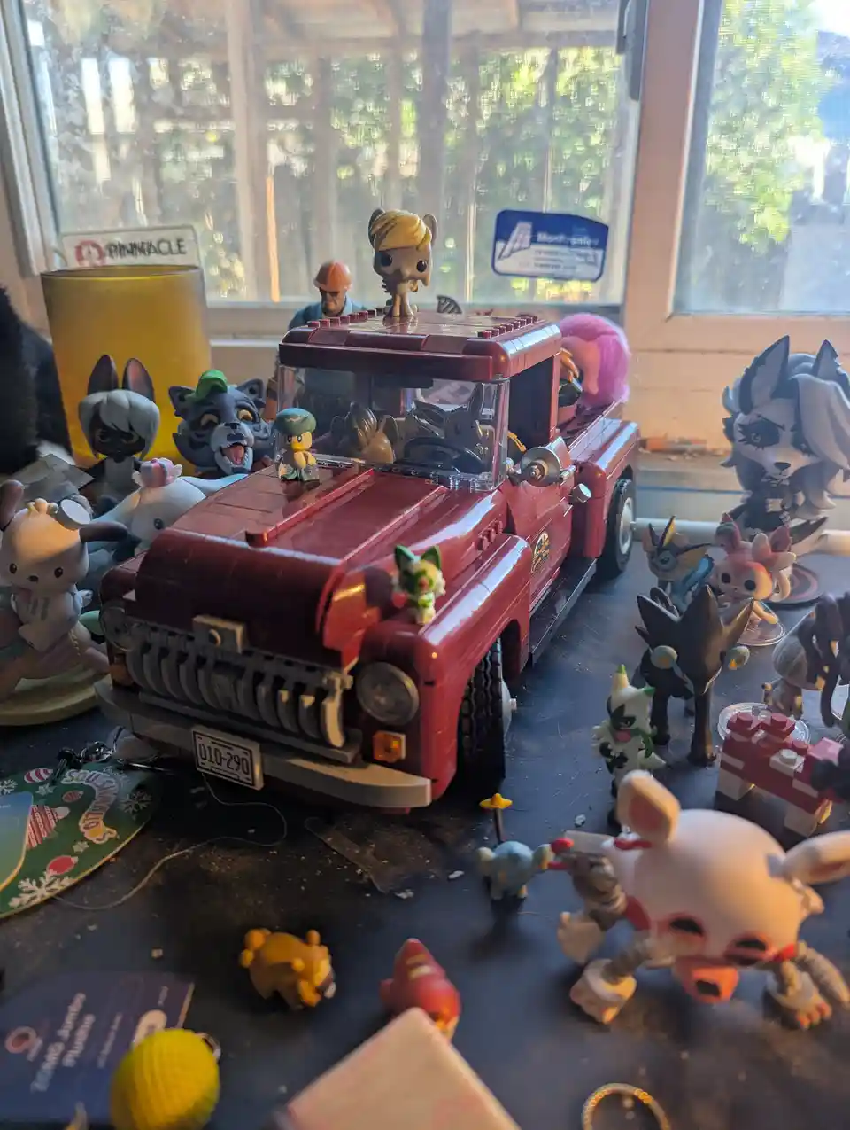 A red toy truck sits on a cluttered table surrounded by numerous small figurines and collectibles, including Funko Pops and…
