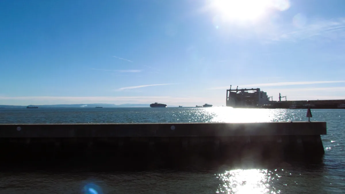 A bright, sunny day at a harbor with a concrete pier in the foreground, ships visible on the water, and a large industrial…