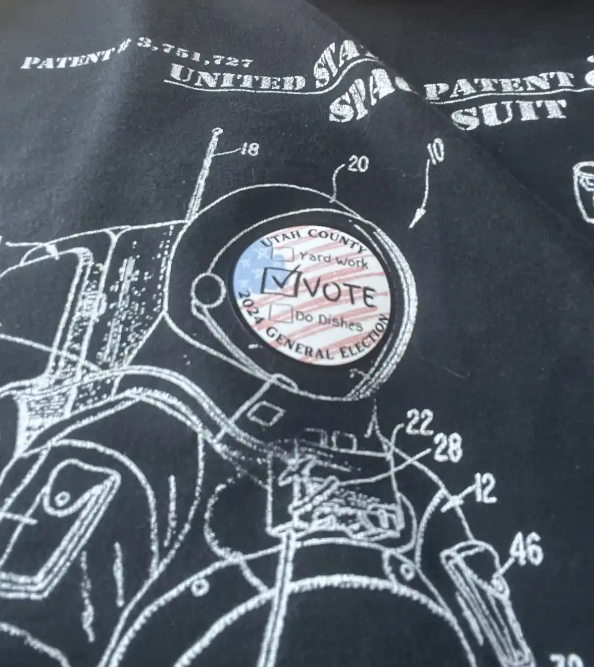 A close-up of a black t-shirt featuring a white line drawing resembling a patent diagram of a spacesuit, labeled 'PATENT…