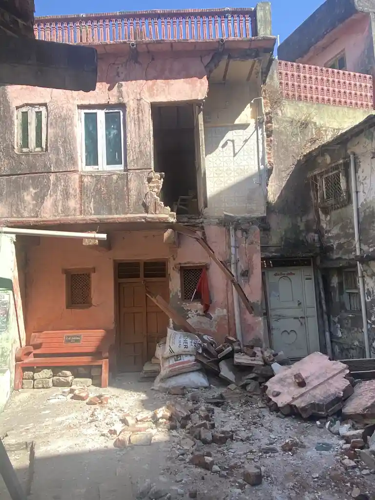 a dilapidated, partially demolished pink house with rubble and debris scattered on the ground, a wooden bench sits intact in…