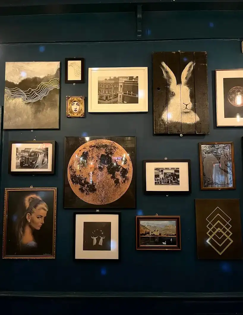 A dark blue wall is covered with numerous framed artworks of various styles and subjects, including a large textured moon, a…