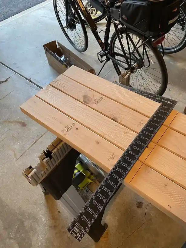 A high-angle photo shows several light-colored wooden planks arranged on a workbench, with a long black ruler laid across…