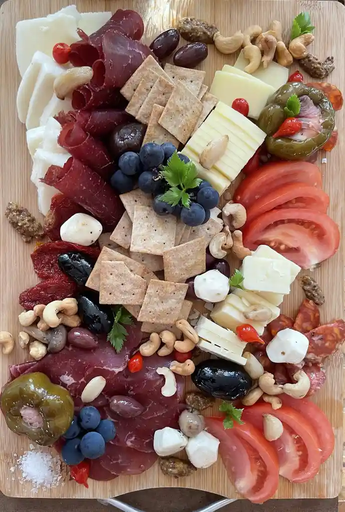 An overhead shot of a wooden cutting board filled with an assortment of food items including sliced meats, various cheeses…