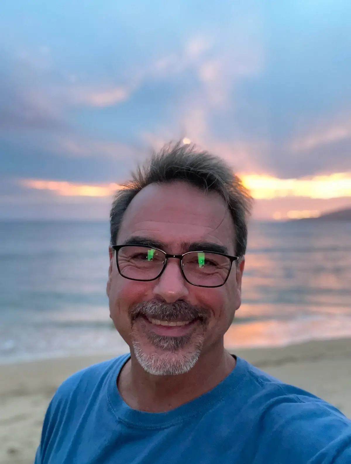 A smiling man with glasses and a blue t-shirt takes a selfie on a beach at sunset. The sky is painted with orange and blue…