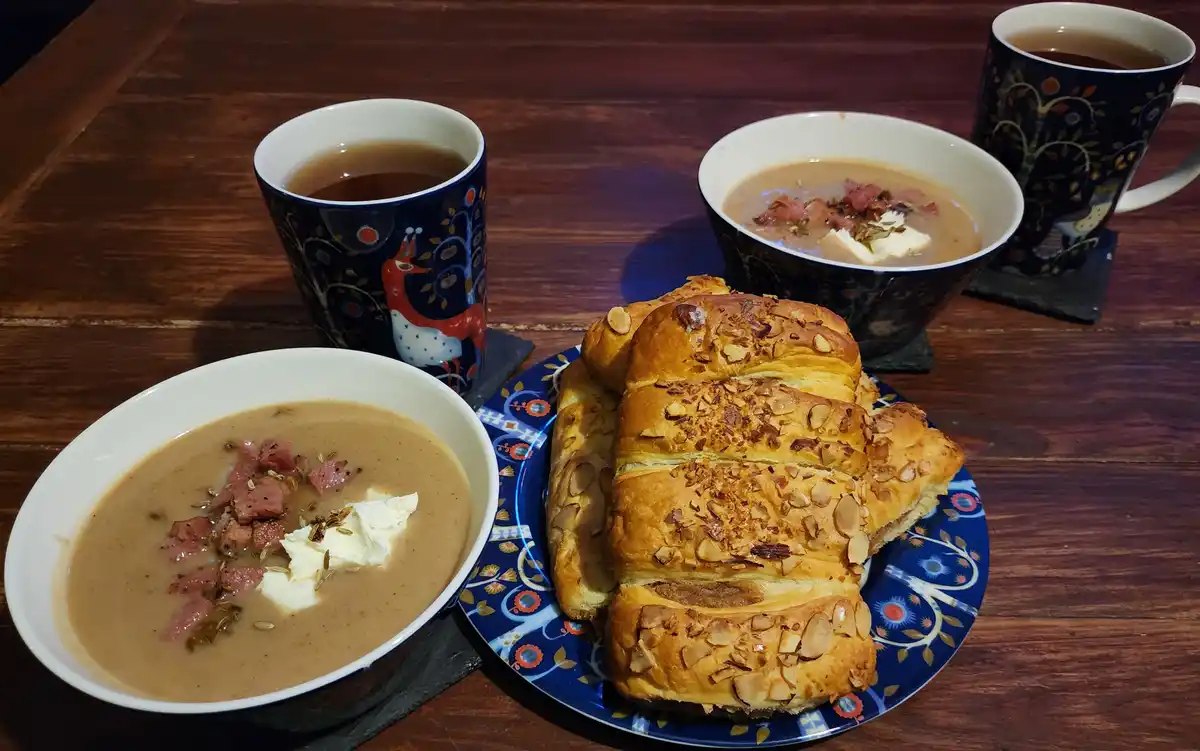 A wooden table displays two bowls of creamy soup topped with diced meat, fennel seeds, and butter, alongside a plate of…
