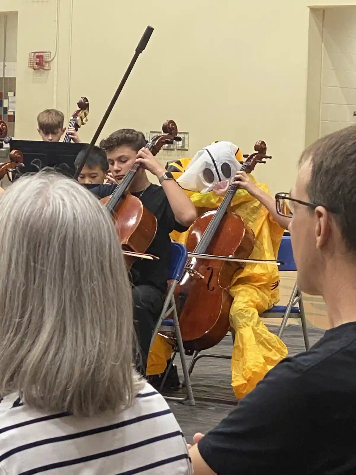 A person in a yellow tiger mascot costume with a white mask plays a cello in an orchestra, surrounded by other musicians and…
