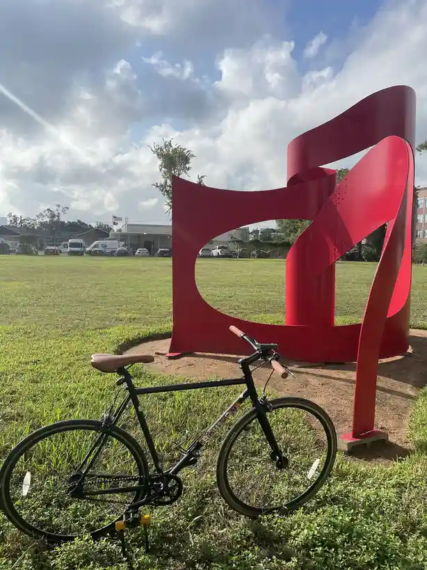 A black bicycle with brown handlebars and seat is parked on green grass next to a large, abstract red metal sculpture shaped…