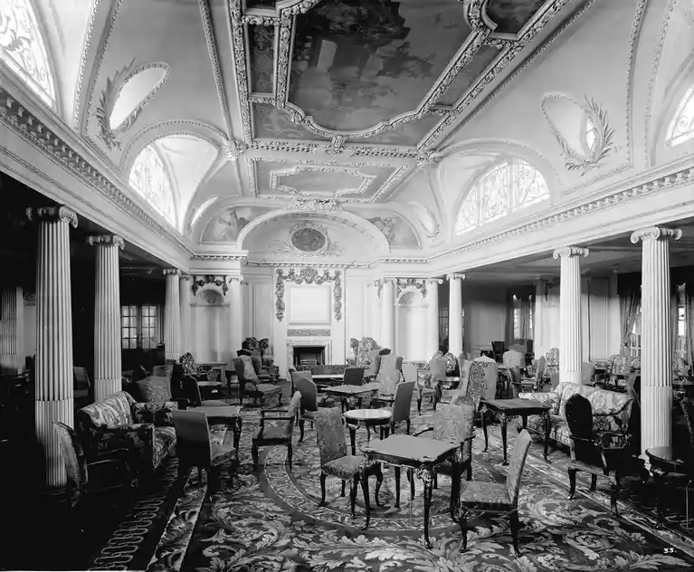 a black-and-white photograph shows an opulent, empty lounge with ornate ceilings, classical columns, and patterned carpets…