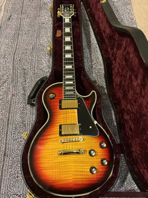 A Gibson electric guitar with a sunburst finish rests inside its open, burgundy velvet-lined case, which sits atop a…