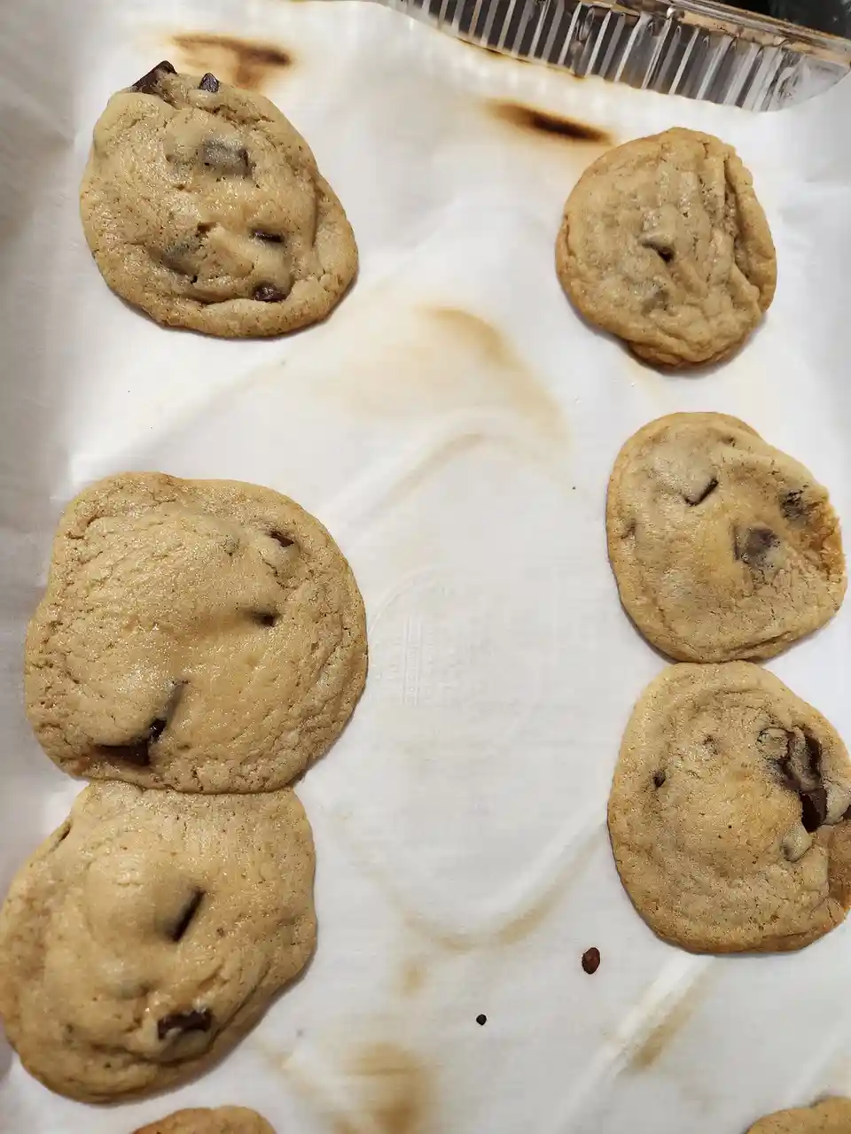 A close-up photo shows six chocolate chip cookies on parchment paper inside a metal baking tray. The cookies vary in…
