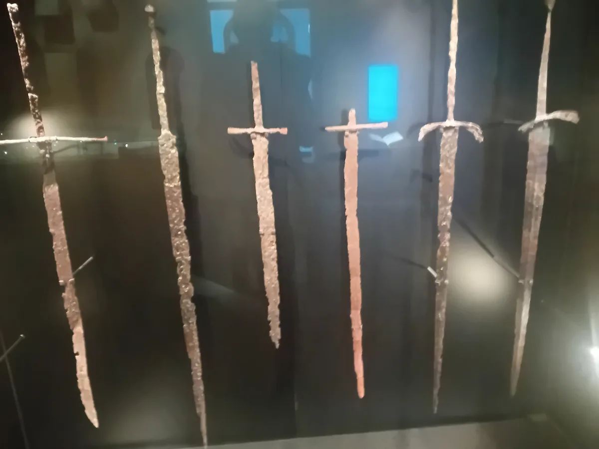 Several heavily corroded, ancient swords are displayed vertically in a dark museum case, their blades and hilts covered in…