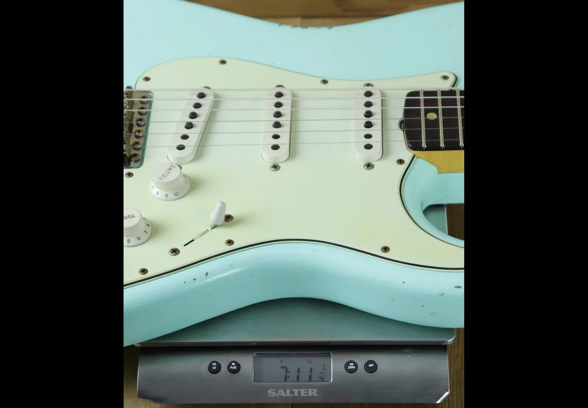 A light blue electric guitar with a cream pickguard is placed on a digital kitchen scale. The scale's display reads '7:114'…