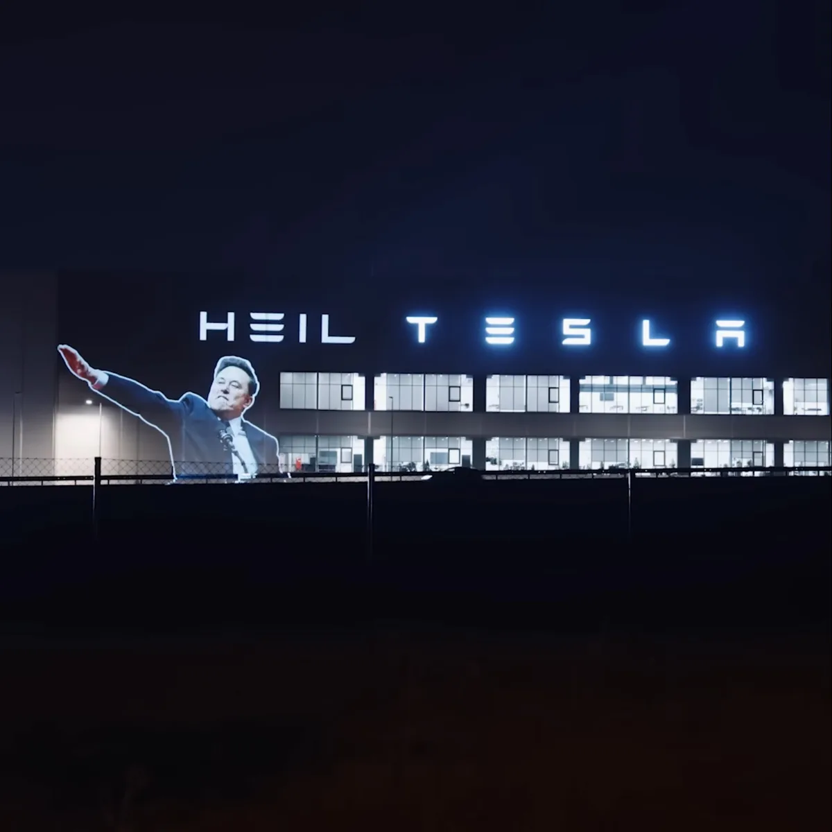 A nighttime photo of a large building with illuminated signage that reads 'HEIL TESLA', where the 'E' in 'TESLA' is replaced…