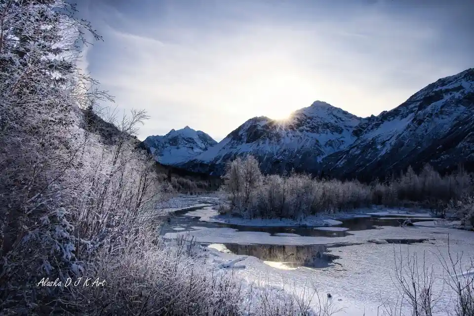 A serene winter landscape photograph shows snow-covered mountains with the sun rising or setting behind them, casting a soft…