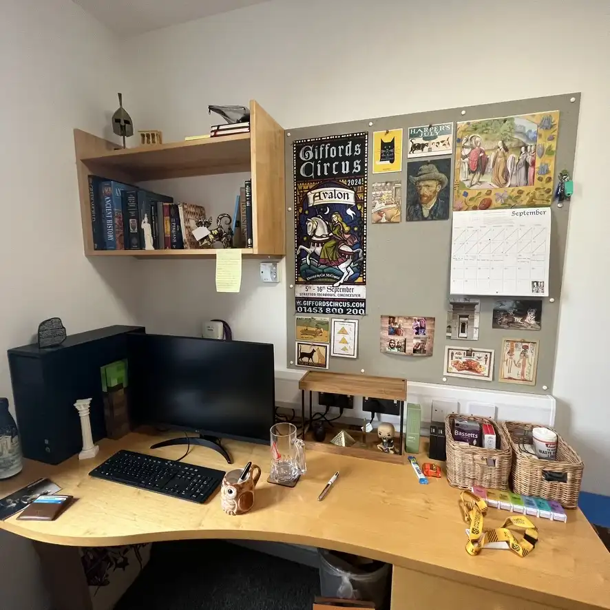 A cluttered home office desk with a computer, keyboard, mug, and various personal items. A corkboard behind the desk is…