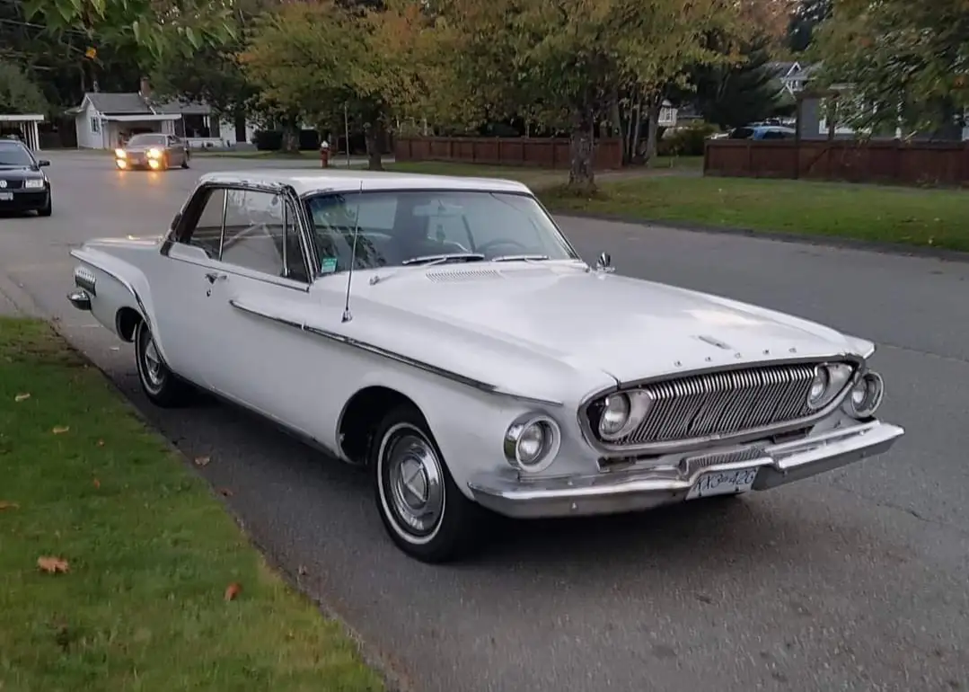 A white 1960s American classic car, likely a Rambler, is parked on a residential street with autumn trees and houses in the…
