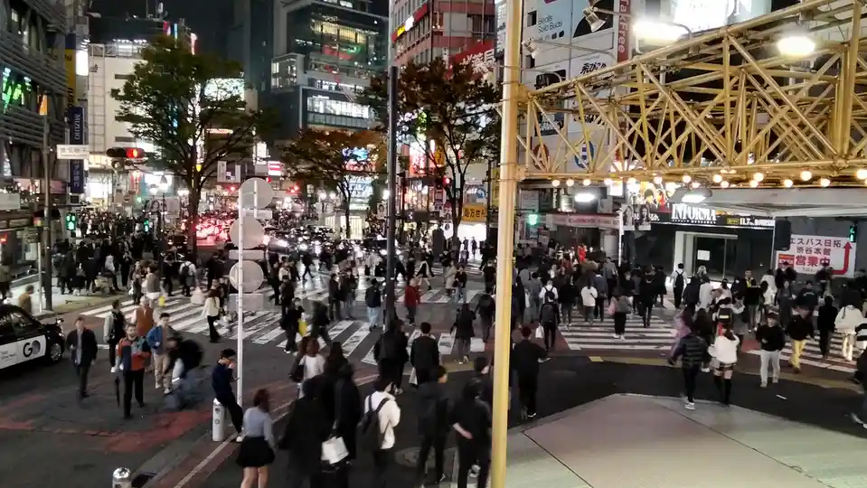 A crowded, bustling Tokyo street intersection at night, filled with pedestrians crossing the road and cars waiting at…
