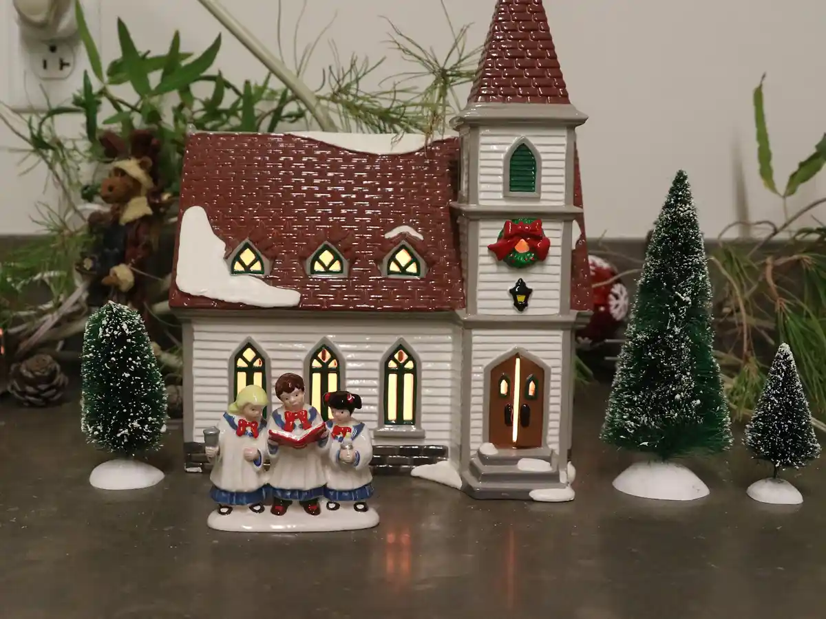 A ceramic Christmas village scene features a lit church with three singing angel figurines in front, surrounded by miniature…