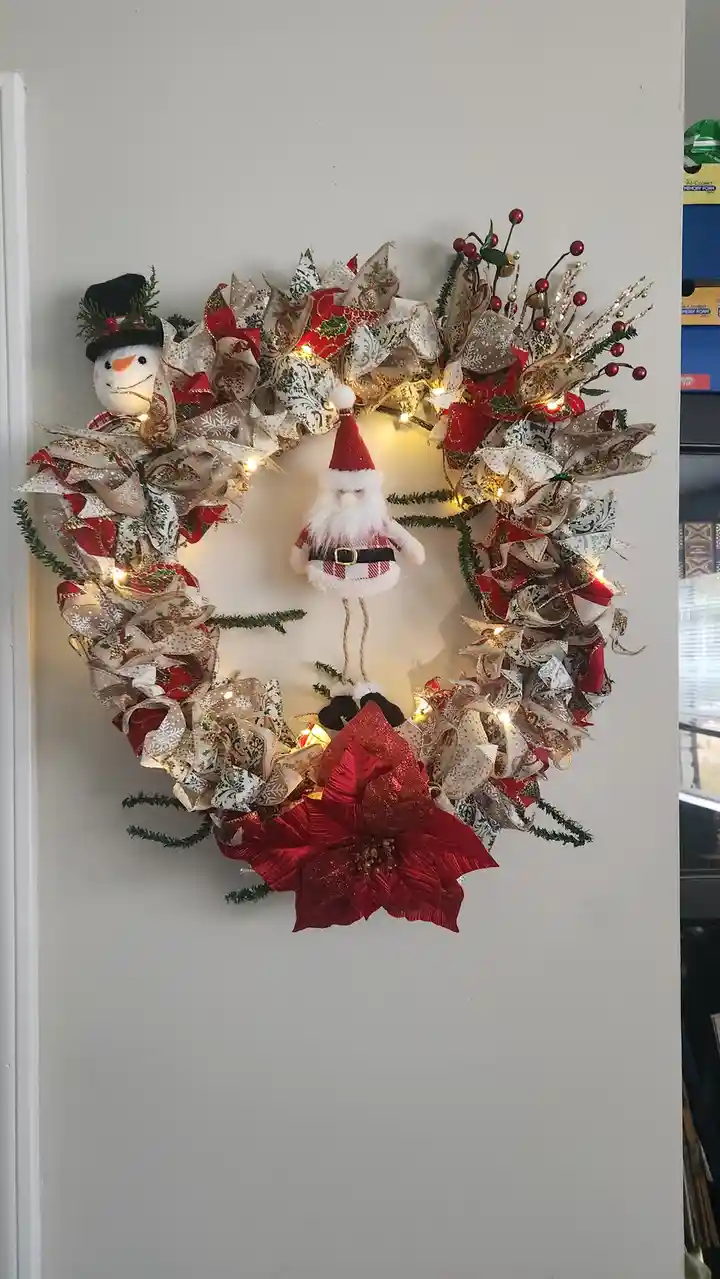 A heavily decorated Christmas wreath with red and gold ribbons, lights, and greenery features a small Santa Claus figurine…