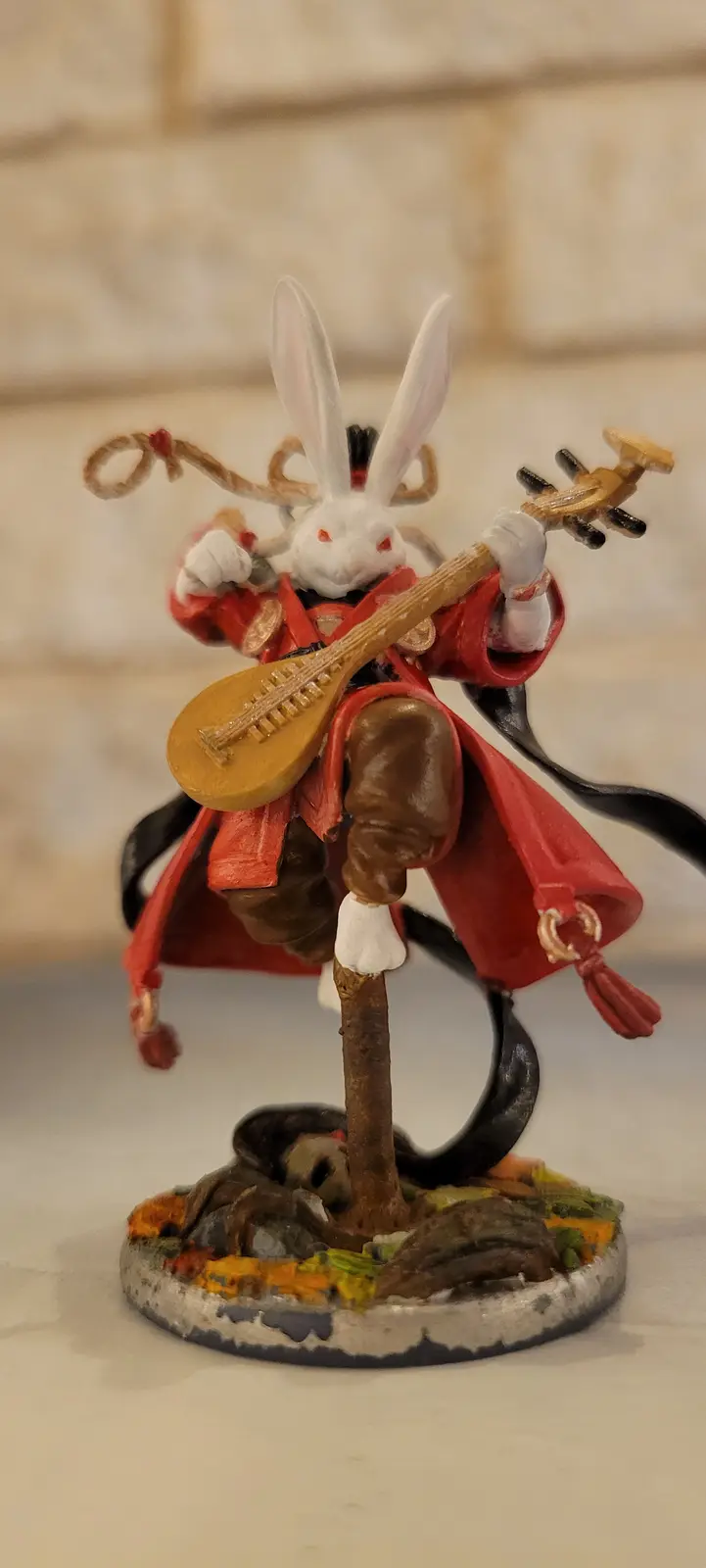 A painted miniature figurine of a white rabbit with red eyes, wearing a red robe and brown pants, is posed dynamically while…