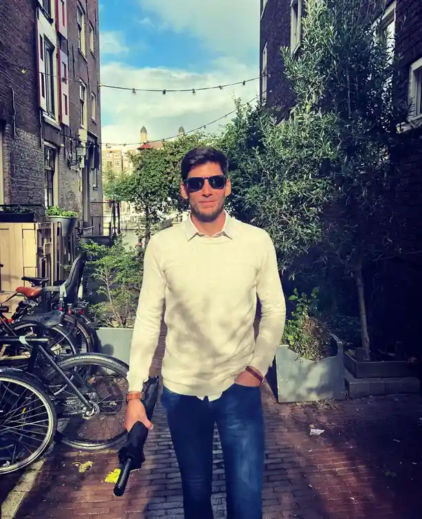 A man wearing sunglasses, a light-colored sweater, and jeans stands on a brick path between buildings, holding a closed…