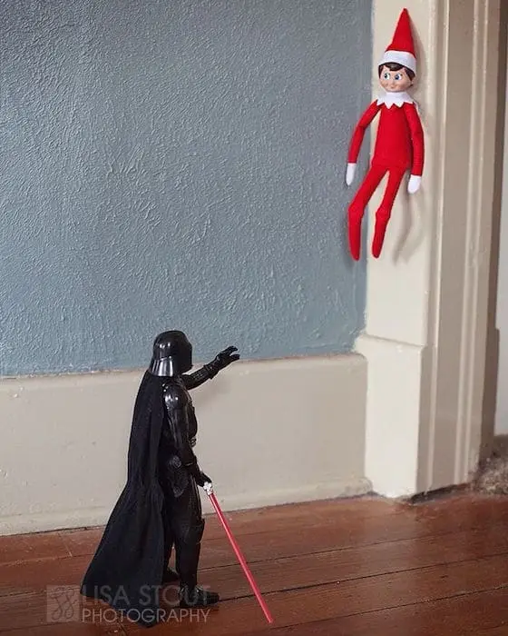 A small red-clad elf on the shelf is perched on a white doorframe, looking down at a Darth Vader action figure holding a red…