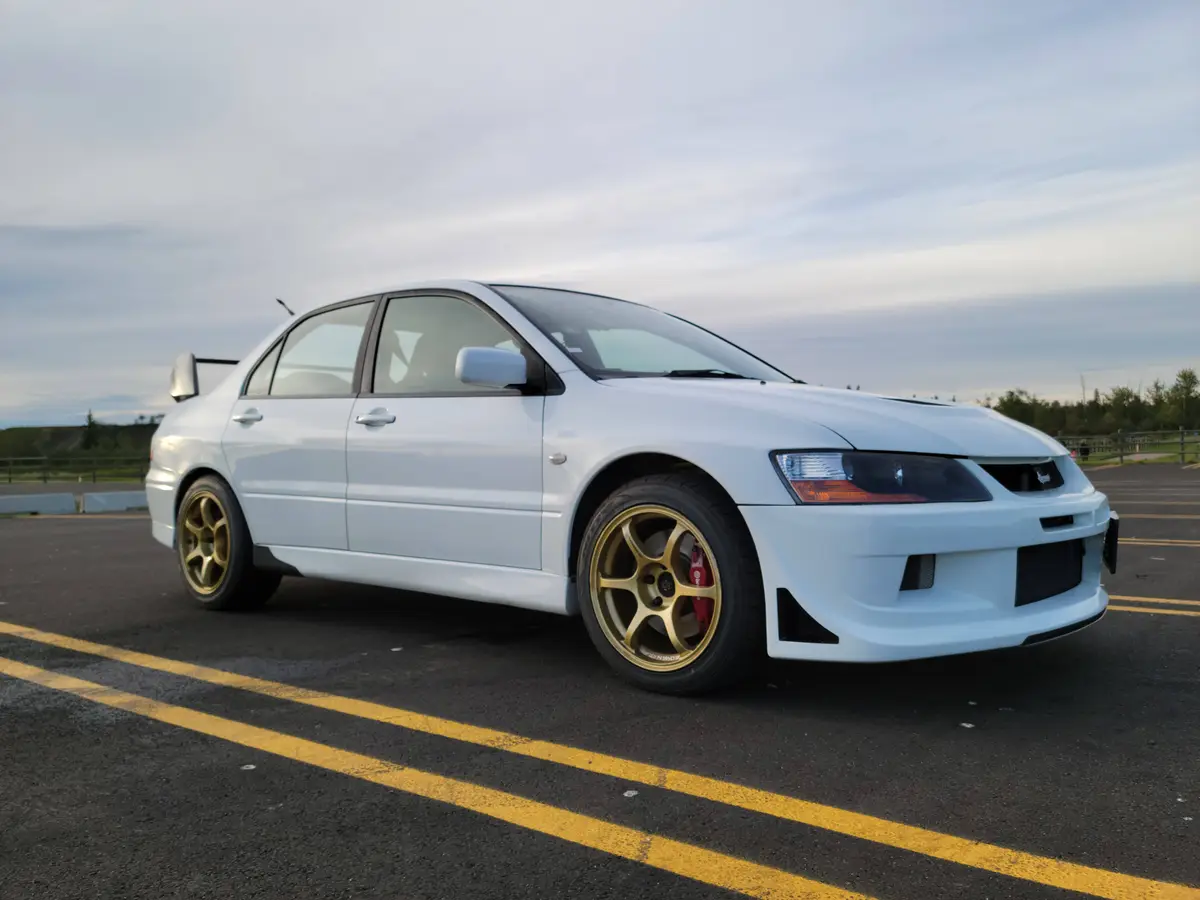 A white Mitsubishi Lancer Evolution sedan is parked in a paved parking lot with yellow lines, featuring gold-colored…