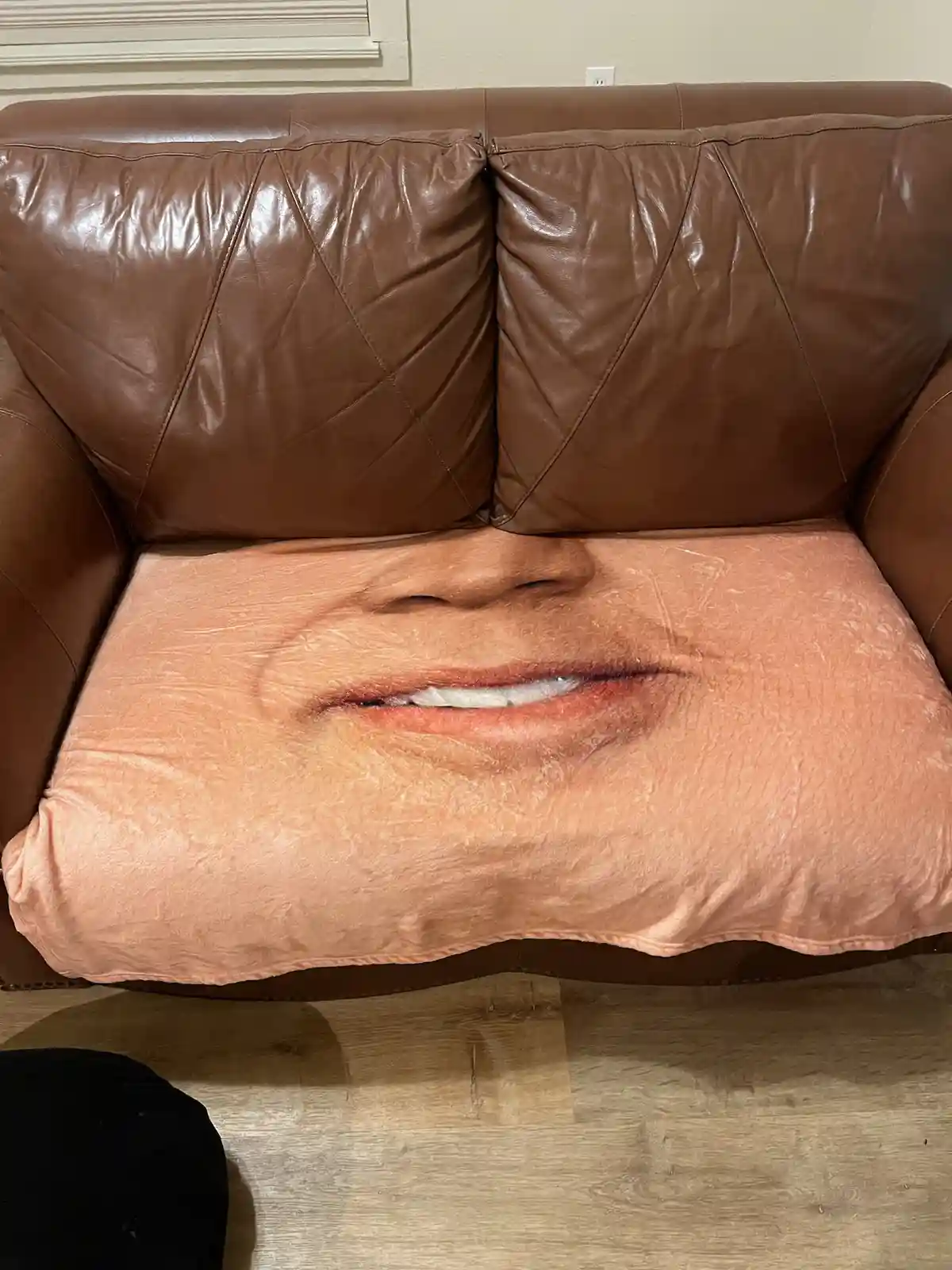 A brown leather couch has a large, printed pillow on its seat that shows a close-up of a smiling human face, specifically…