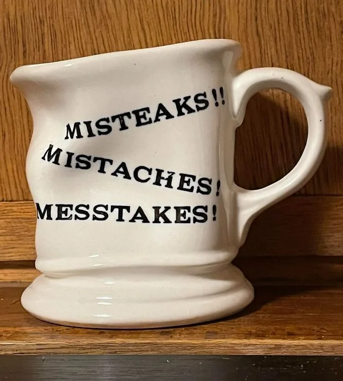 A white ceramic mug with a handle sits on a wooden surface, featuring the text 'MISTEAKS!! MISTACHES! MESSTAKES!' printed in…
