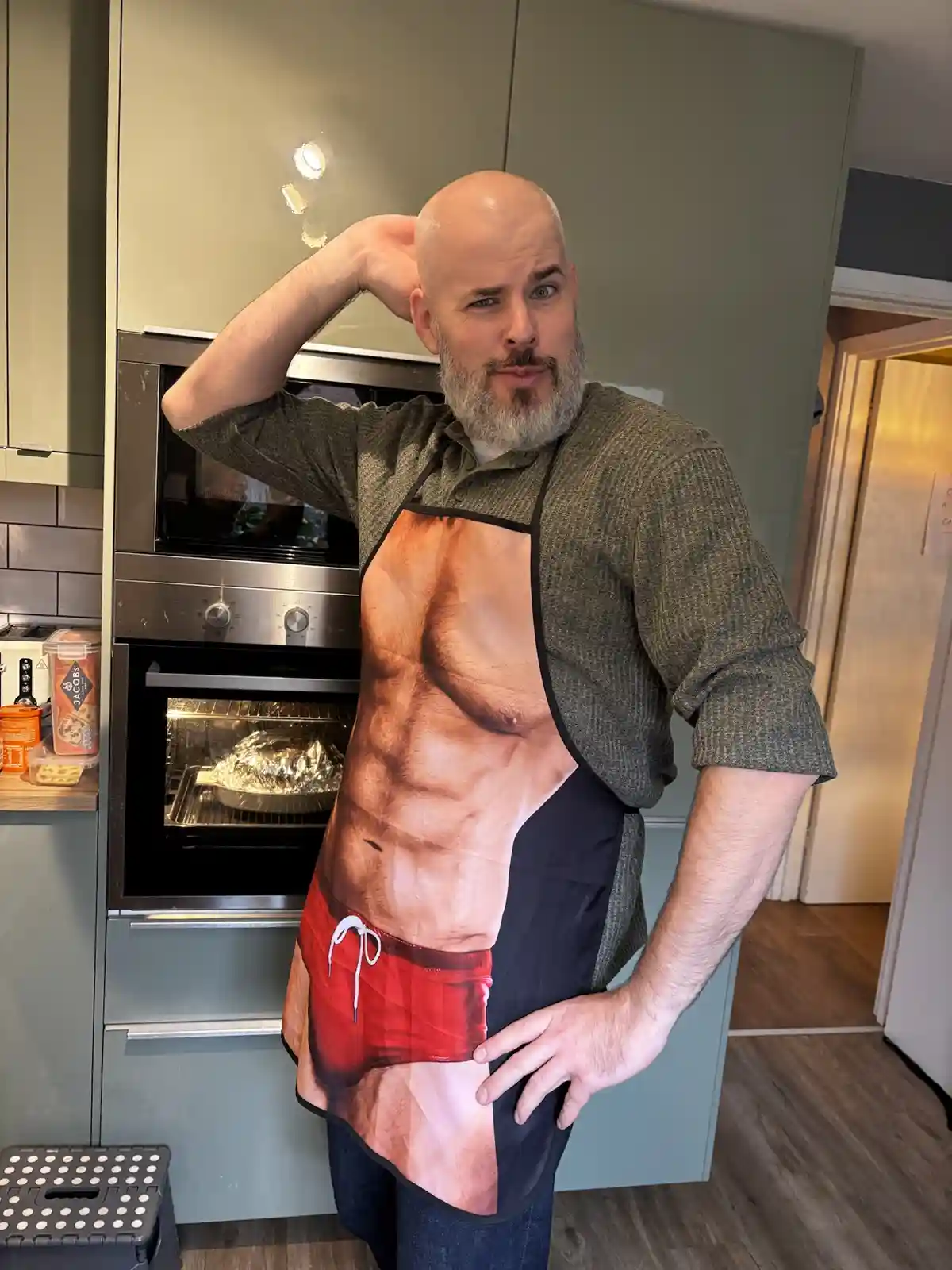 A bald man with a beard stands in a kitchen wearing an apron that visually depicts a muscular male torso and red swim…