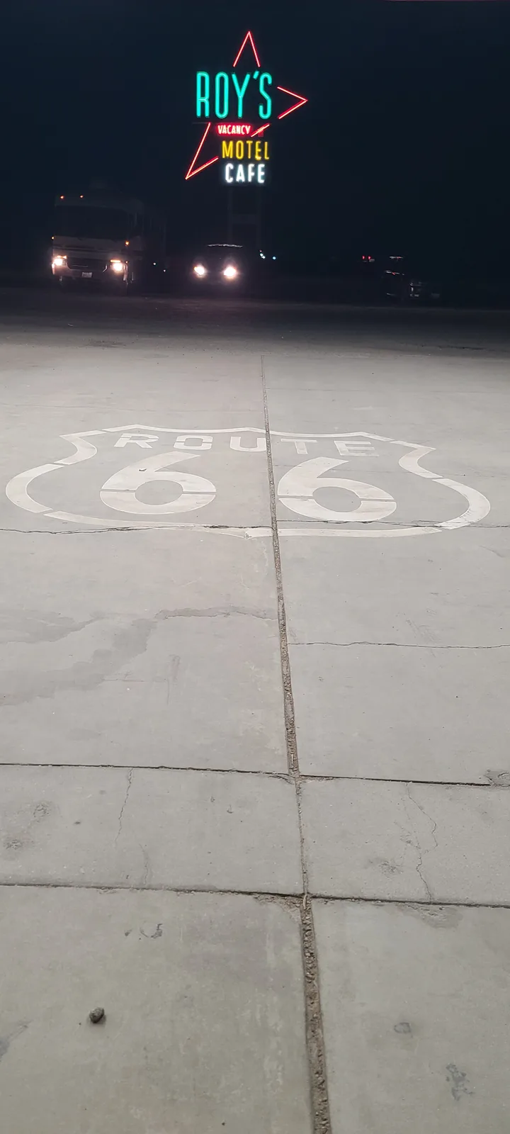 A low-angle night photo shows a cracked concrete surface with a faded white Route 66 shield painted on it, leading toward a…