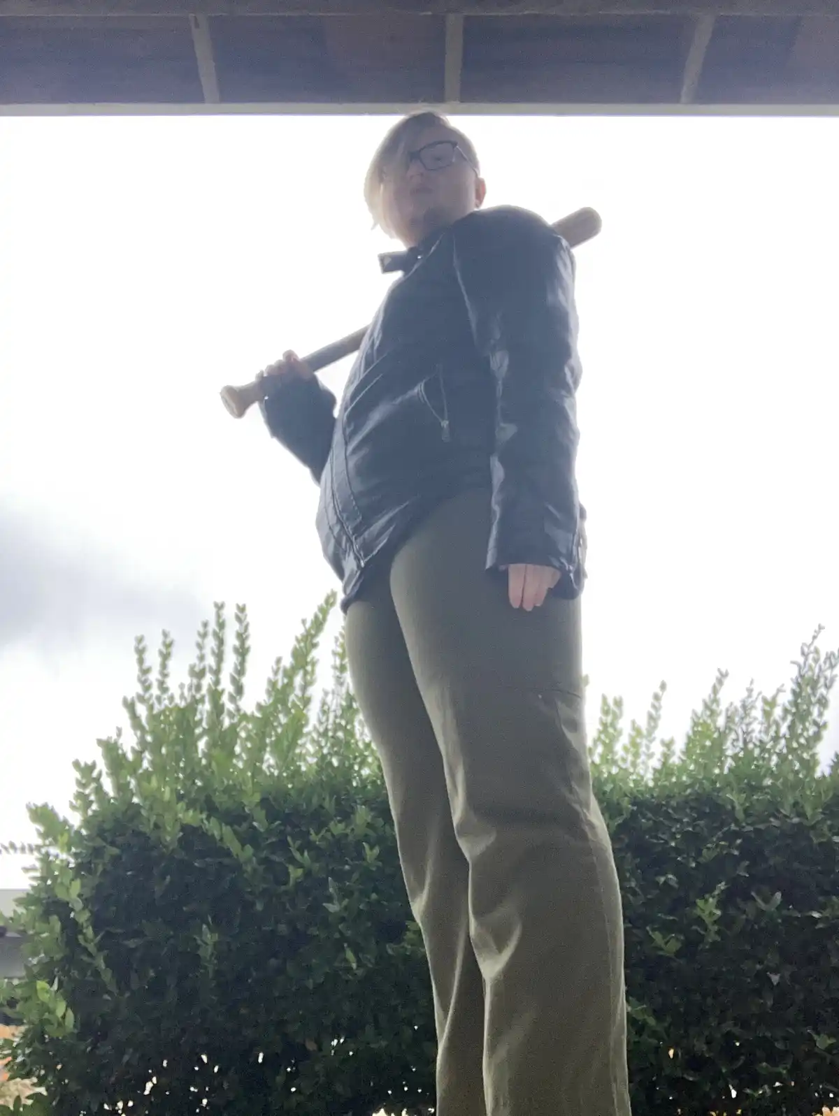 A person with short hair and glasses, wearing a black jacket and olive pants, stands outdoors under a covered porch holding…
