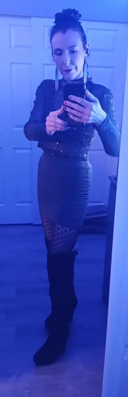 A woman stands in front of a mirror in a dimly lit room bathed in blue light, taking a selfie with her phone. She wears a…