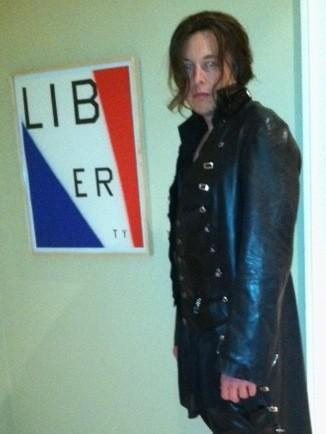 A person with shoulder-length brown hair and glasses stands in a black leather coat next to a framed poster. The poster…