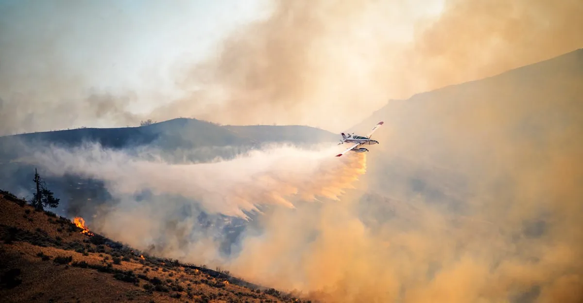 A red-and-white firefighting aircraft flies low over a smoky, fire-ravaged hillside, releasing a stream of water or fire…