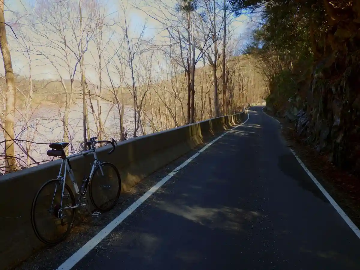 A silver road bicycle leans against a concrete barrier on a winding, tree-lined road next to a river, with the overlaid text…