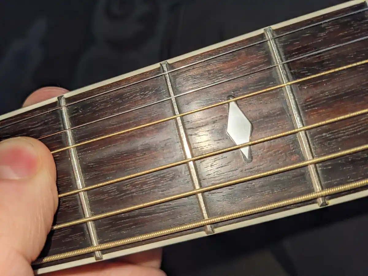 A close-up photo shows a person's thumb pressing down on the fretboard of a dark wood guitar, with the metal frets and…