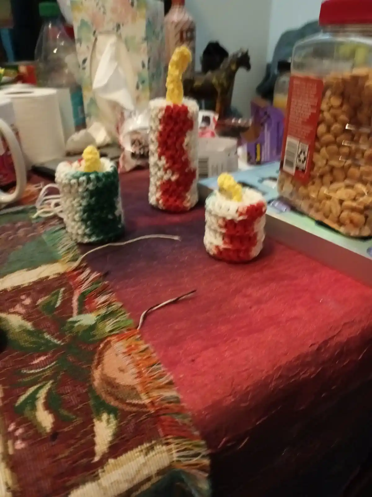 Three small, hand-crocheted candles in red-and-white, green-and-white, and red-and-white stripes sit on a red and patterned…