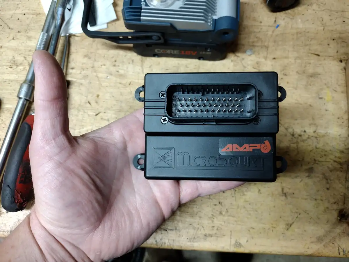 A hand holds a small black AMP'd MicroSquirt electronic control unit with a multi-pin connector, set against a cluttered…