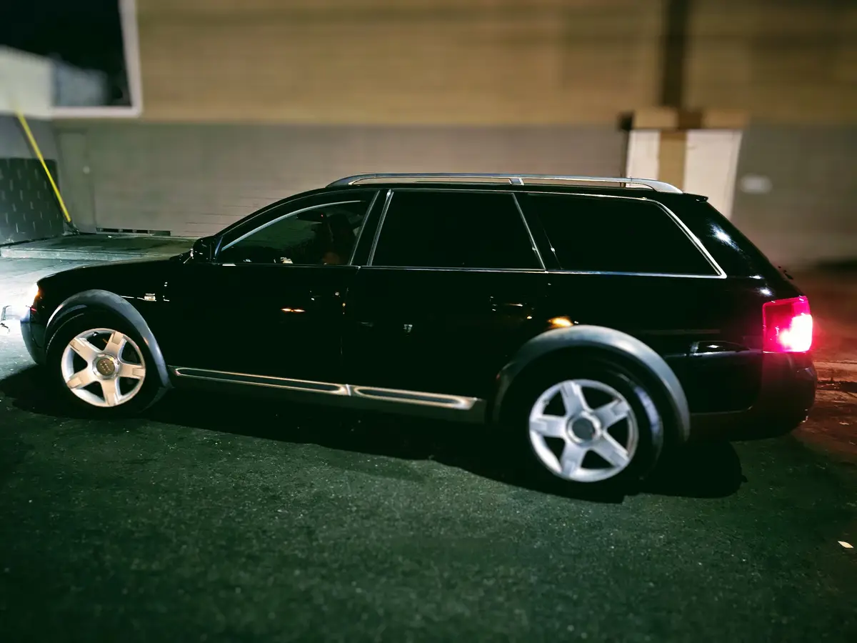 A black station wagon, likely an Audi Allroad, is parked at night with its taillight on, showing a glossy finish and silver…