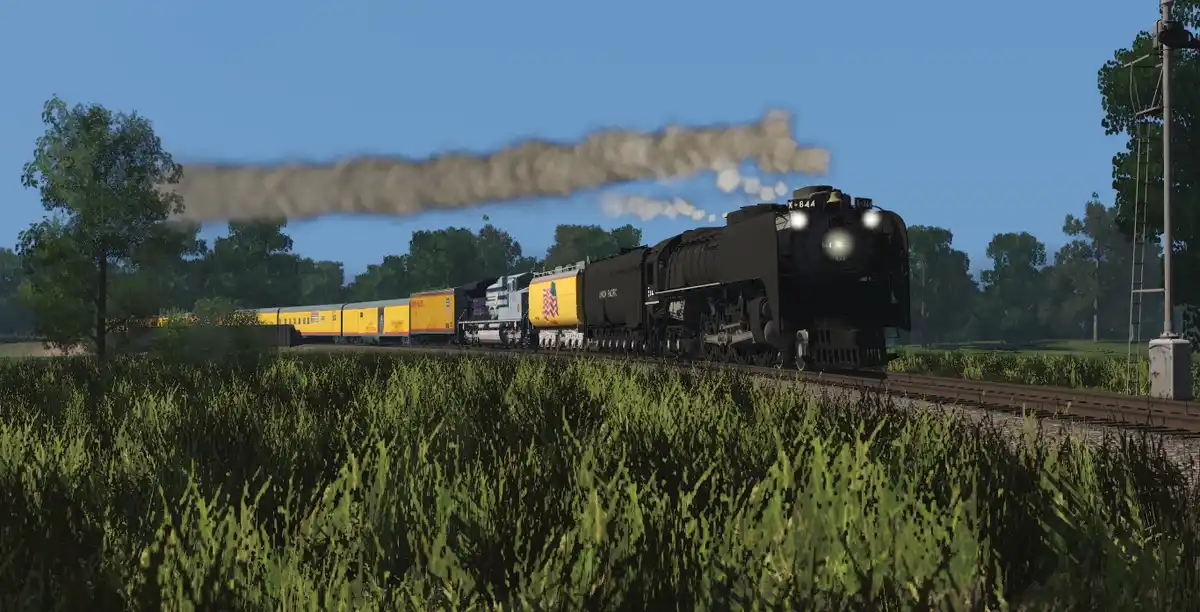A steam locomotive pulling a long train through a field of tall green grass under a clear blue sky, with smoke billowing…