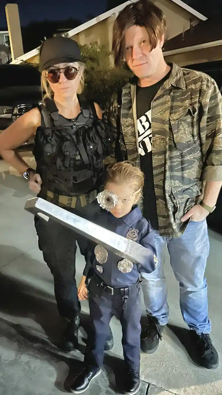A family of three poses outdoors at night: a woman in tactical gear and sunglasses, a man in a camouflage shirt and wig, and…