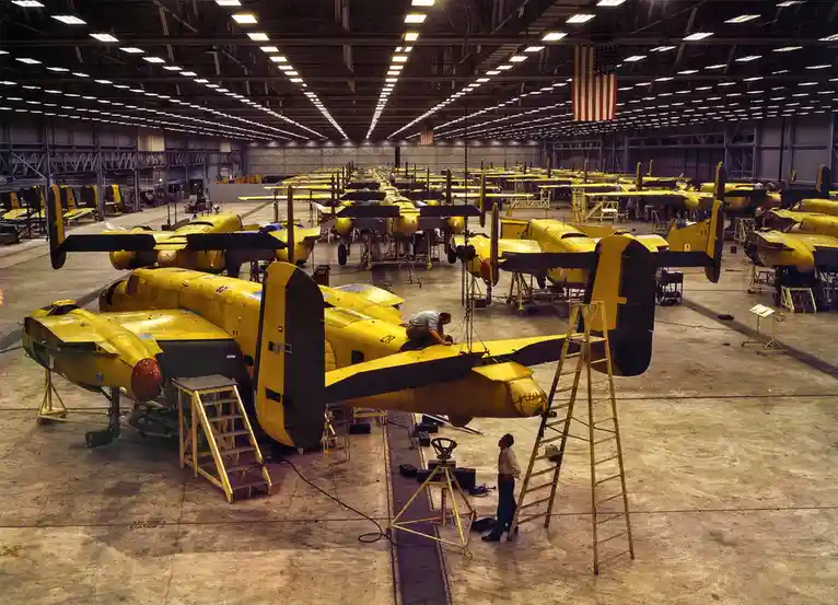 Rows of yellow military aircraft fuselages are being assembled in a massive hangar, with workers on ladders and scaffolding…
