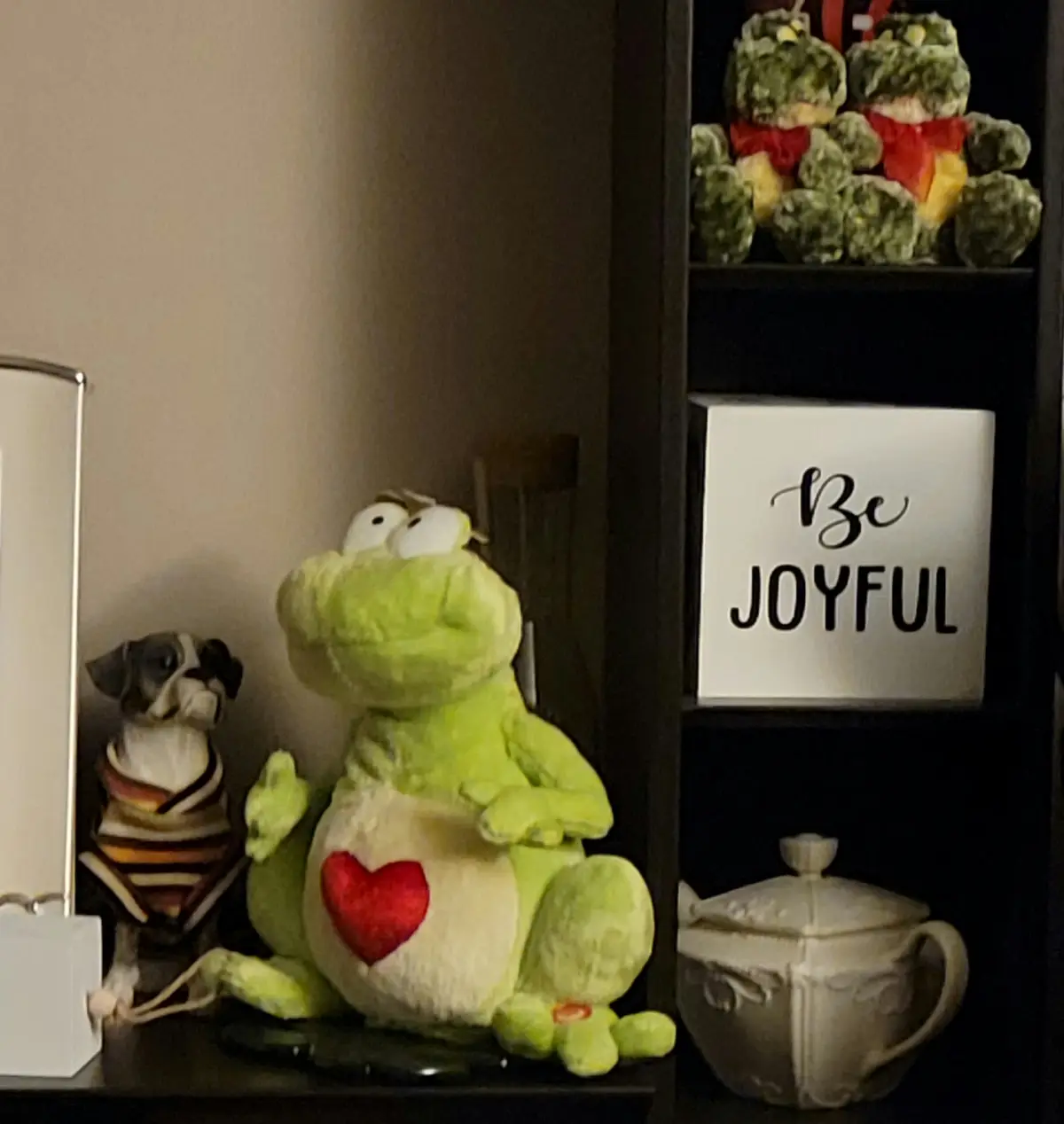 A green plush frog with a red heart on its belly sits on a shelf next to a small dog figurine wearing a striped sweater…
