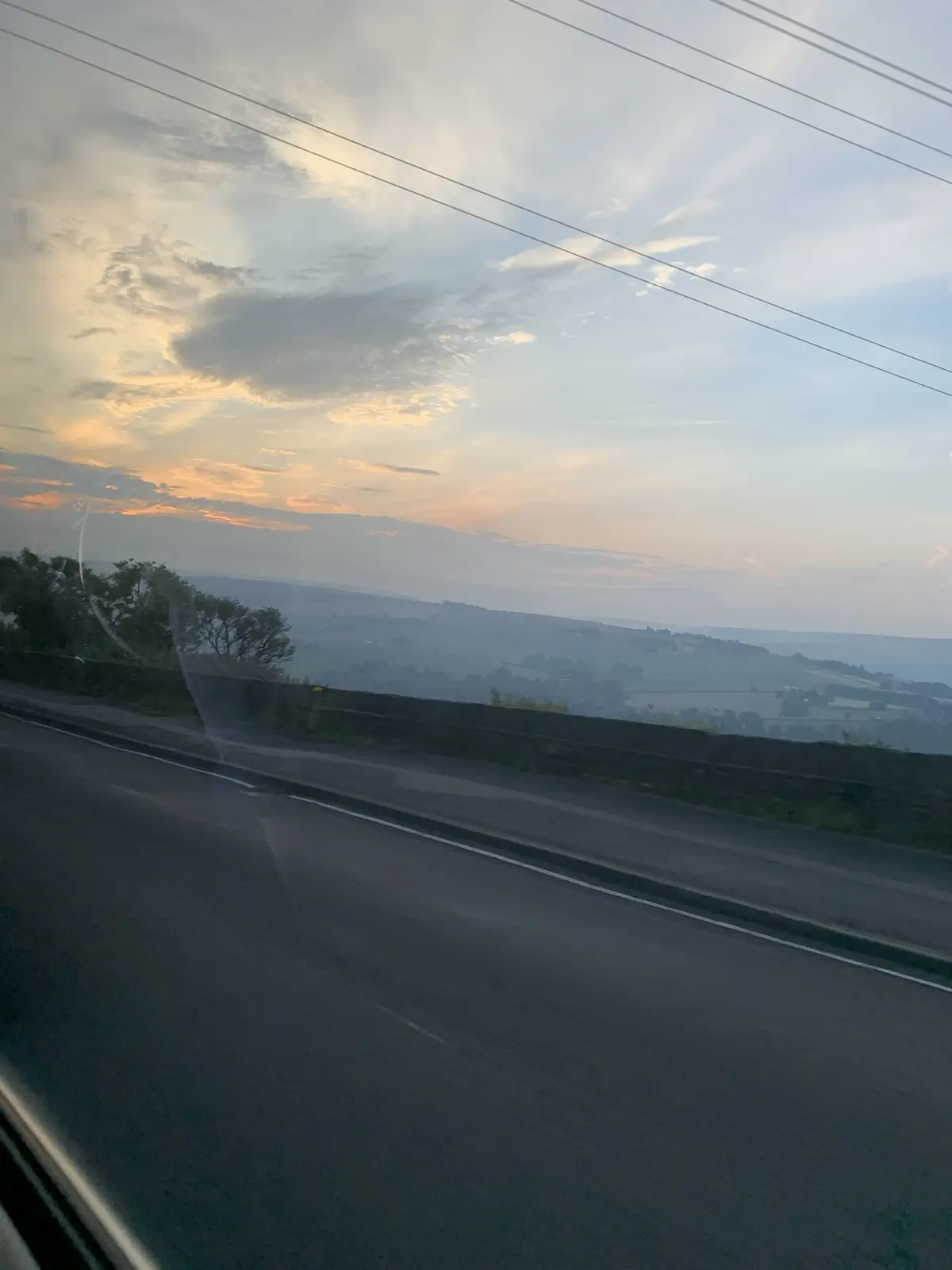 A photo taken from inside a car looking out at a scenic countryside road during sunset or sunrise, with rolling hills…