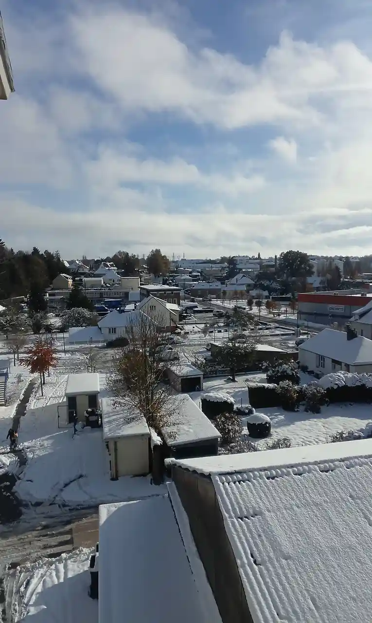A high-angle photo shows a residential neighborhood blanketed in snow under a partly cloudy sky, with rooftops, trees, and…