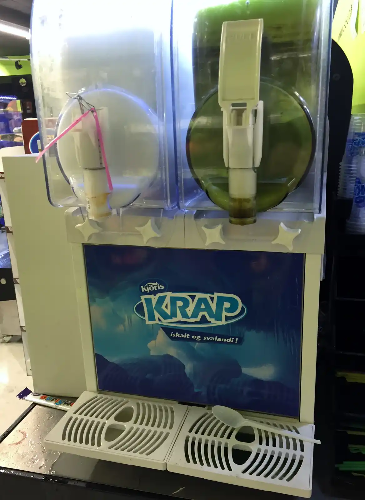 A commercial slushie machine branded 'Kjoris KRAP' with two dispensers, one containing a clear liquid and the other a green…