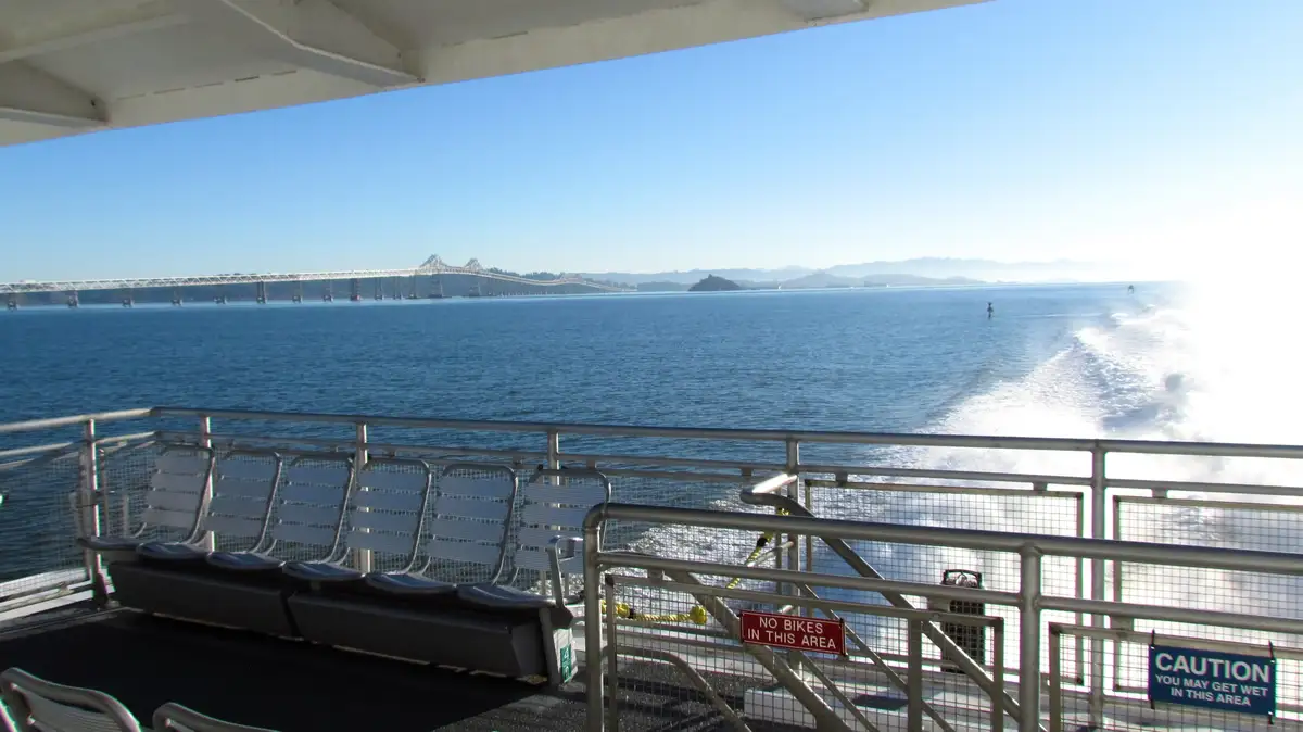 A photo taken from the deck of a moving ferry shows a scenic view of a long bridge over blue water under a clear sky. In the…